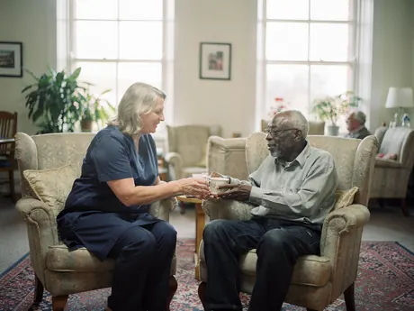 A care worker passing a cup of tea to an elderly resident in a care home lounge
