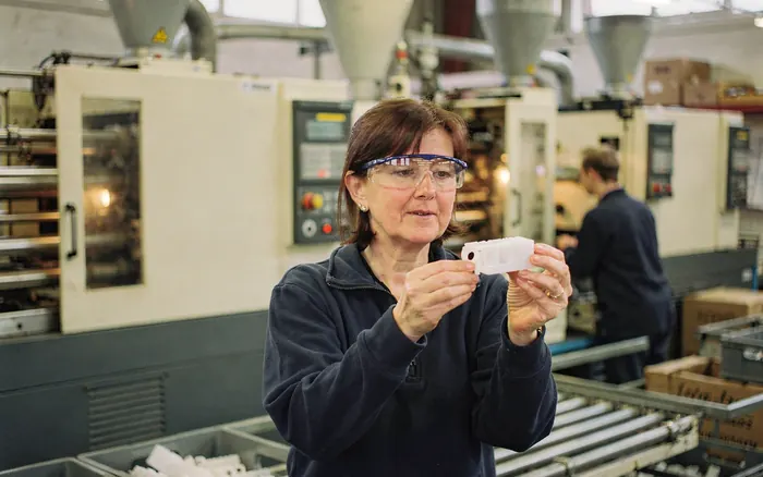 A factory owner checking a moulded part beside energy-intensive injection moulding machines