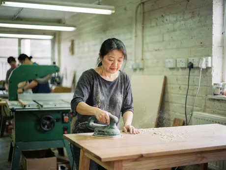 A woodworker sanding a tabletop in a busy furniture workshop